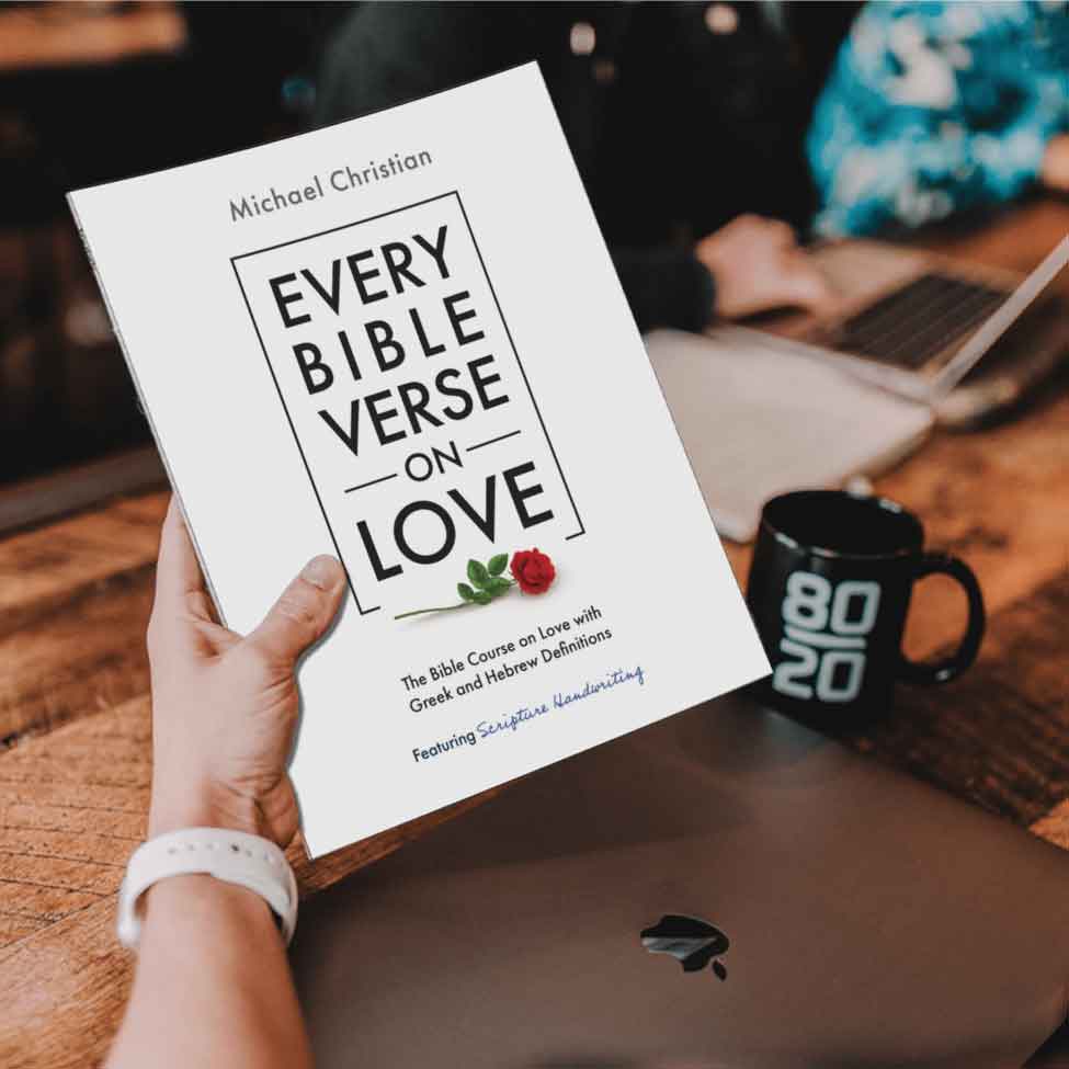 Image of a man's hand holding the book Every Bible Verse on Love over a laptop computer with a coffee cup on the side